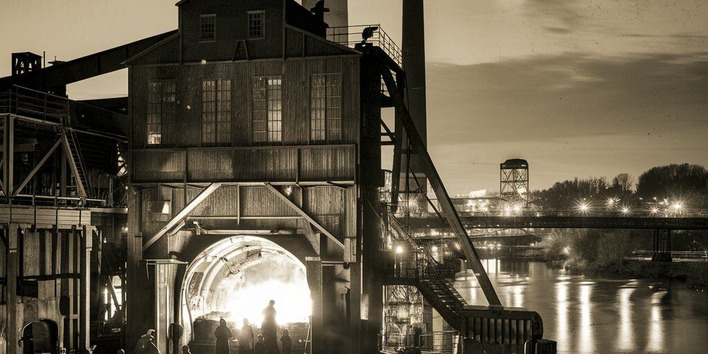 A gritty, sepia-toned illustration of a 19th-century Pittsburgh iron furnace roaring to life at night, workers silhouetted against the molten glow, with the Monongahela River in the background.