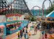 A vibrant, nostalgic scene of Kennywood Park in Pittsburgh, Pennsylvania. The image features classic wooden roller coasters like the Jack Rabbit and Thunderbolt, colorful vintage carnival rides, and the famous Potato Patch fries stand. Families walk through the park on a sunny summer day, with kids holding balloons and riding the merry-go-round. The background shows the iconic Monongahela River view, and the atmosphere is cheerful, warm, and timeless, capturing over 125 years of amusement park history.