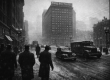 Black and white winter street scene in downtown Pittsburgh in the late 1940s with the Fort Pitt Hotel towering above Penn Avenue, vintage cars and pedestrians in coats and fedoras, and light snow in the air.