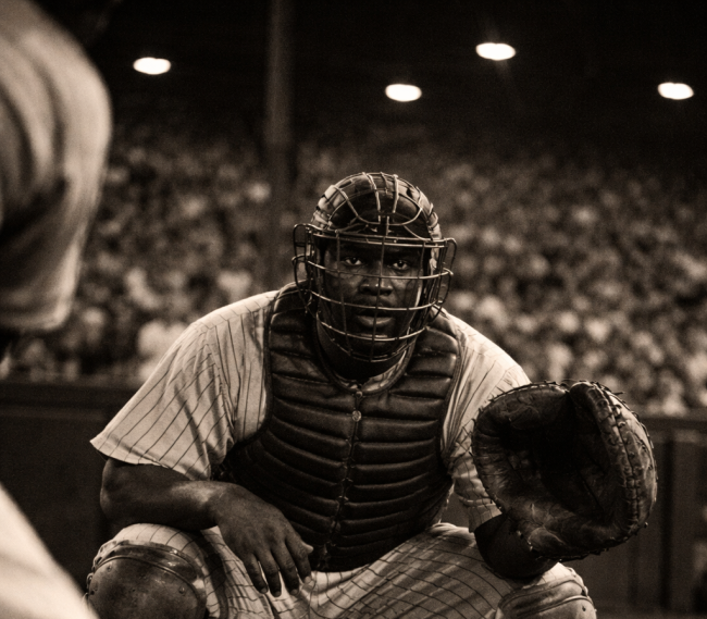 Baseball catcher in 1940s uniform crouches behind home plate at a packed Pittsburgh stadium, capturing the era when Josh Gibson was considered the greatest hitter in baseball.