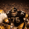 A Pittsburgh Steelers defensive lineman in the classic 1970s black and gold uniform breaks through an offensive line under stadium lights, capturing the intensity of the Steel Curtain defense.