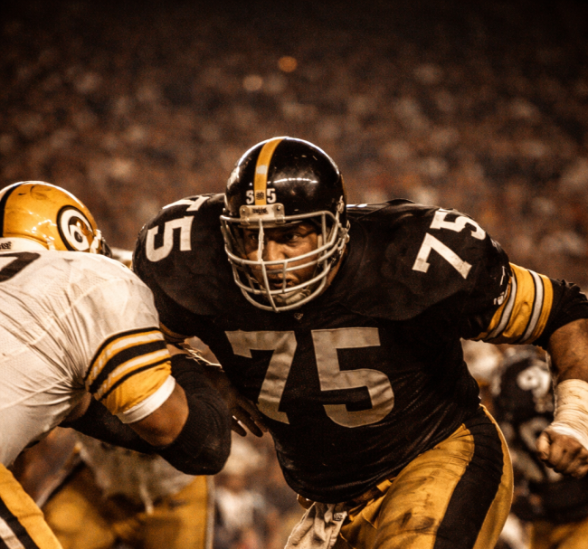 A Pittsburgh Steelers defensive lineman in the classic 1970s black and gold uniform breaks through an offensive line under stadium lights, capturing the intensity of the Steel Curtain defense.